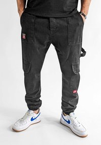 Schwarze Cargo-Hosen mit elastischen Bündchen, ausgestattet mit mehreren Taschen und einem kleinen roten Logo. Kombiniert mit weißen Sneakers mit blauen Akzenten.