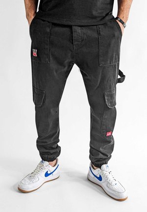 Black cargo pants with elastic cuffs, featuring multiple pockets and a small red logo. Paired with white sneakers with blue accents.