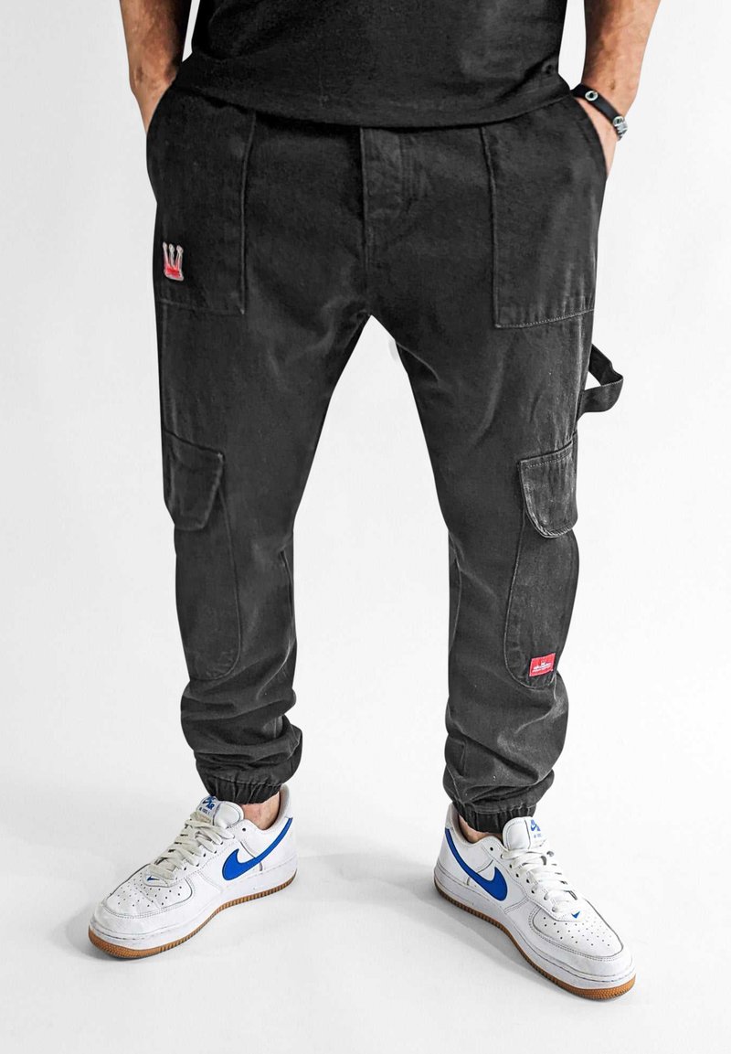 Schwarze Cargo-Hosen mit elastischen Bündchen, ausgestattet mit mehreren Taschen und einem kleinen roten Logo. Kombiniert mit weißen Sneakers mit blauen Akzenten.