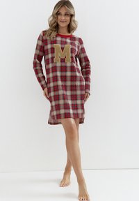 Czerwona i beżowa nocna sukienka w kratę z długimi rękawami, okrągłym dekoltem i dużą teksturowaną literą "M" na przodzie. Posiada rozcięcia po bokach u dołu.