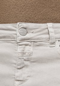 Un jean en denim blanc présente un bouton circulaire argenté portant l'inscription "CLOSED", avec des coutures visibles et une texture subtile dans le tissu.