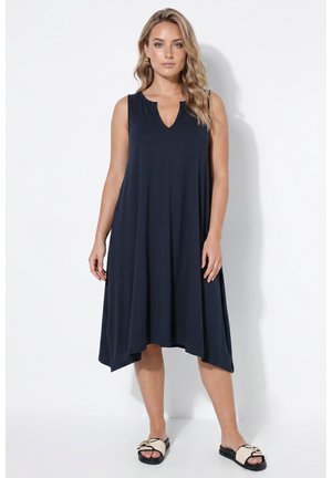 Femme aux cheveux blonds ondulés portant une robe bleu marine sans manches jusqu'aux genoux et des sandales tissées noires et blanches, debout sur fond blanc.