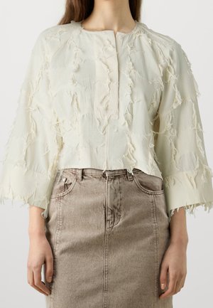 Femme portant un chemisier à manches longues texturé crème avec des détails effilochés, associé à une jupe en jean beige taille haute.