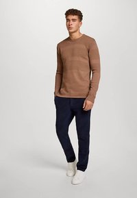Brauner, strukturierter, langärmeliger Strickpullover mit rundem Ausschnitt, kombiniert mit dunkelblauen Hosen und weißen Turnschuhen. Einfaches Design, figurbetonter Stil.