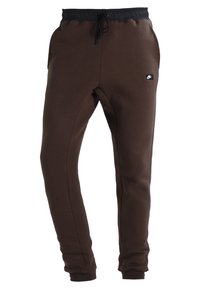 Pantalones de chándal marrones hechos de tejido suave, con una cinturilla de cordón negra y bolsillos laterales. Pequeño logotipo negro en la parte delantera.