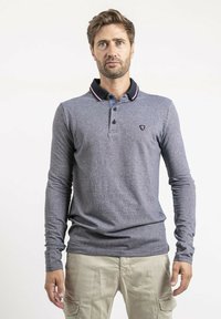 Polo à manches longues bleu marine en tissu texturé, avec un col noir contrastant orné de détails rouges et un petit emblème sur la poitrine.