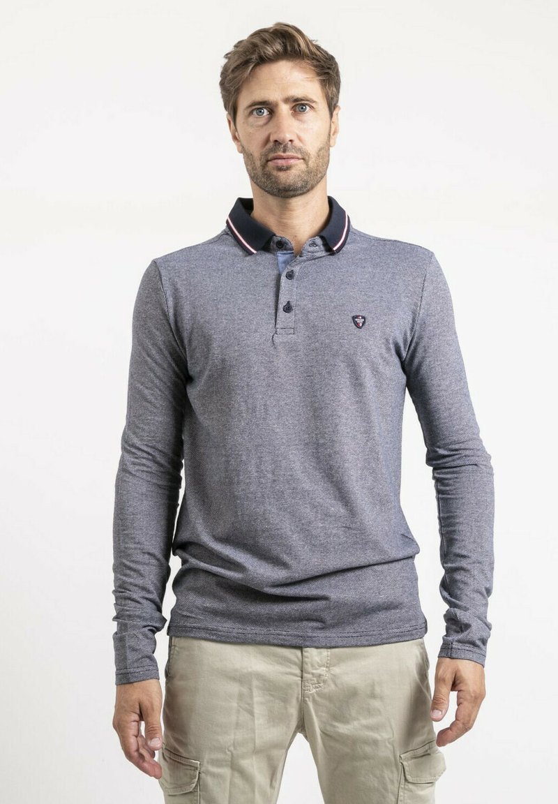 Polo à manches longues bleu marine en tissu texturé, avec un col noir contrastant orné de détails rouges et un petit emblème sur la poitrine.