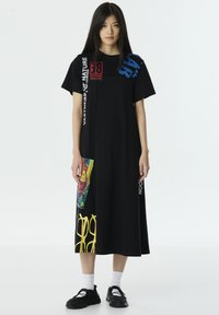 Zwart katoenen t-shirtjurk met korte mouwen, een relaxte pasvorm, kleurrijke grafische accenten en tekstdetails in rood, blauw en geel.