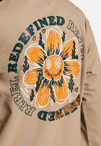 Beige sweatshirt med et stort orange blomstermotiv med sorte konturer, omgivet af teksten "REDEFINED REBEL STREETWEAR" i grøn.
