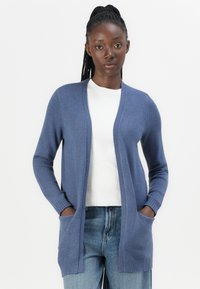 Långärmad blå cardigan i mjukt material, med öppen framsida, två framfickor och ribbade ärmslut. Bärs över en vit topp.