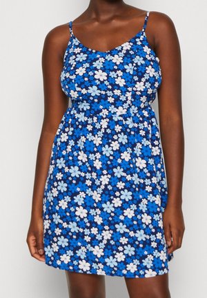 Mujer vestida con un vestido sin mangas de flores azules y tirantes finos, con una cintura ajustada, frente a un fondo liso.