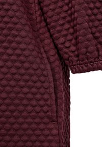 Mörk röd texturerad tyg med ett tredimensionellt diamantmönster, med en lös passform och elastiska ärmslut för flexibilitet.