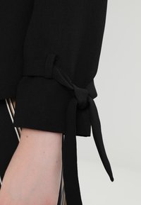 Černá blůza s manžetou a vázáním, která má jemnou texturu a strukturovaný design. Látka je hladká a má čistý, minimalistický vzhled.