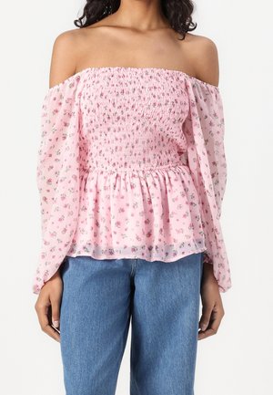 Woman wearing light pink off-shoulder blouse with floral print and smocked bodice, paired with blue denim jeans, standing against white background.