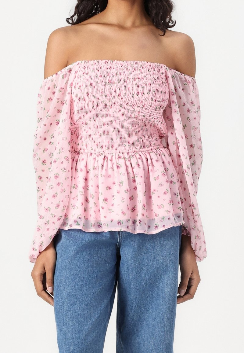 Femme portant un chemisier rose pâle à épaules dénudées avec imprimé floral et corsage smocké, associé à un jean bleu, debout devant un fond blanc.