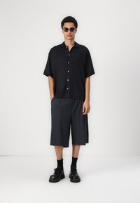 Svart t-skjorte med kort erme og knappestenging, kombinert med mørkegrå knelange shorts; modellen har på seg solbriller og kraftige svarte støvler.