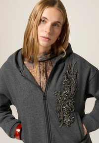 Felpa grigia con zip, decorata con intricati ricami floreali di perline sul fronte, abbinata a una leggera blusa floreale sotto.