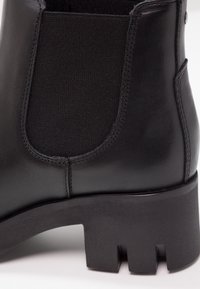Botín negro de cuero hasta el tobillo con paneles laterales elásticos y un tacón grueso y texturizado en negro. Superficie lisa con detalles de costura precisos.