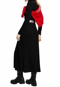 Schwarzes langärmeliges Kleid mit fließender Silhouette, kombiniert mit einem roten gestrickten Sweater, der lässig über die Schultern gelegt ist. Schwarze klobige Stiefel bis zum Knie.