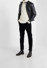 Giubbotto di pelle nera, jumper cremoso a trecce, pantaloni in velluto a coste neri e stivaletti neri alla caviglia. Design aderente, materiali testurizzati e polsini arrotolati.