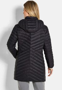 Vrouw in een lange zwarte gevoerde jas met capuchon en chevron-stiksel, met de rug naar de camera, staande tegen een lichtgrijze achtergrond.