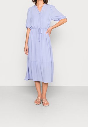 Kobieta ubrana w jasnofioletową sukienkę midi z bufkami na rękawach i paskiem w talii, do tego białe sandały na paski.