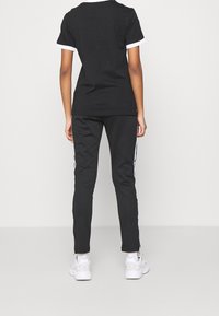 Černé tričko s bílým lemem, spárováno s černými kalhotami s bílými bočními pruhy. Model stojí zády k nám a ukazuje zadní design.