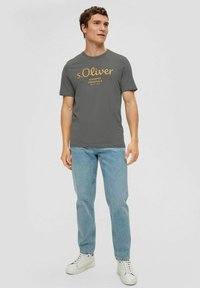 Dark gray cotton t-shirt featuring a light brown logo print, paired with light blue jeans and white sneakers. Simplistic design and fit.