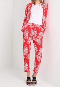 Vrouw in een rood bloemenpak met bijpassende blazer en broek, witte t-shirt en zwarte open teen hakken, staand met handen in de zakken.