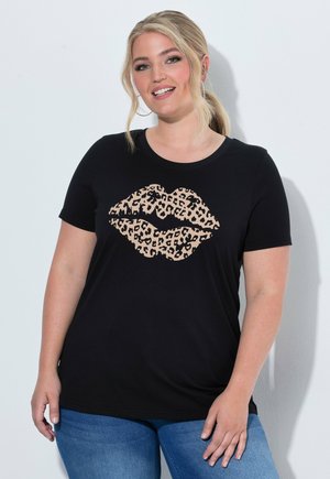 Lächelnde Frau mit blondem Zopf trägt ein schwarzes T-Shirt mit Leopardenlippen-Design und blaue Jeans, steht vor weißem Hintergrund.