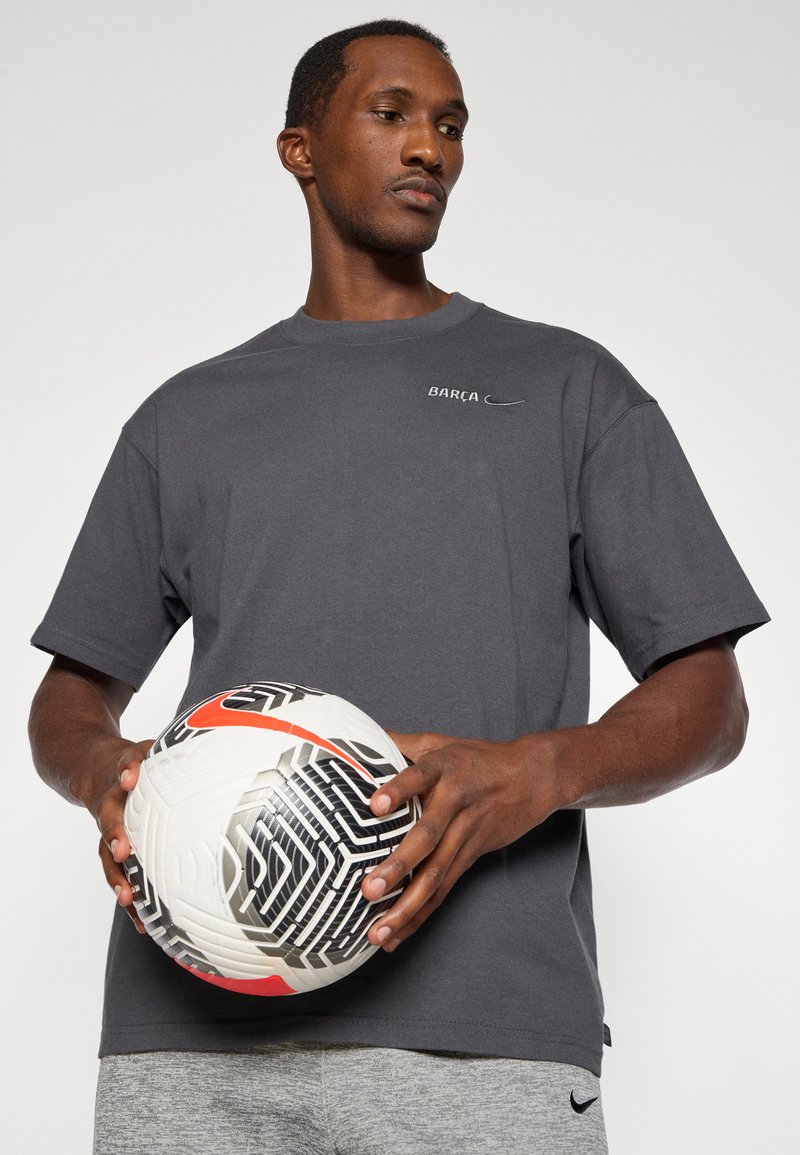 Gray short-sleeve t-shirt with "BARCA" logo in white, paired with gray shorts. Model holds a black and white patterned soccer ball with orange accent.