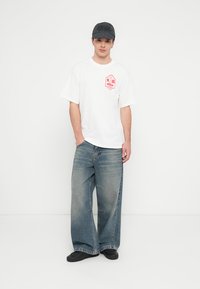 Bílé tričko s krátkým rukávem a červenou grafikou, volné světle modré džíny a černé nazouvací boty. Outfit doplňuje černá čepice.