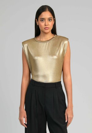 Femme aux longs cheveux noirs portant un haut métallisé doré sans manches et un pantalon noir taille haute, debout devant un fond gris.