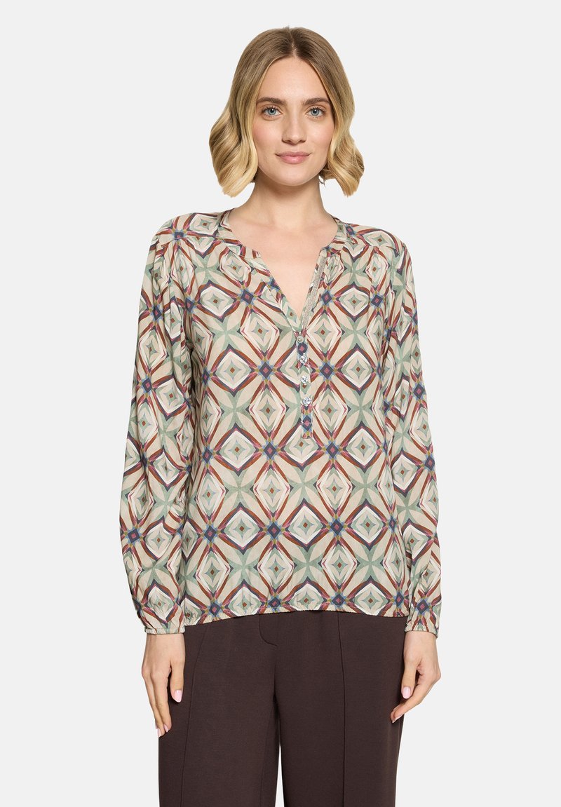 Femme aux cheveux blonds courts portant un chemisier à manches longues aux motifs multicolores et un pantalon sombre, debout devant un fond uni.