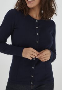 Cardigan blu navy realizzato in morbido tessuto a maglia, con chiusura a bottoni frontale composta da sei bottoni in metallo, maniche a coste e un design aderente.
