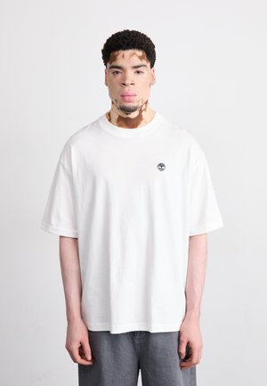 White short-sleeve t-shirt made of cotton, featuring a small blue logo on the left chest and relaxed fit, paired with grey trousers.
