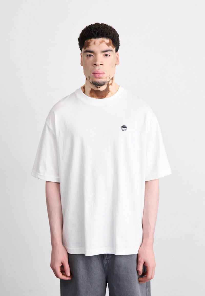 Camiseta blanca de manga corta hecha de algodón, con un pequeño logotipo azul en el lado izquierdo del pecho y un corte relajado, combinada con pantalones grises.