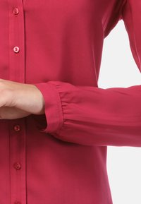 Korte mouwen, knoopshirt van levendig roze stof. Heeft een gladde textuur, een geplooide manchetdetail en bijpassende knopen.