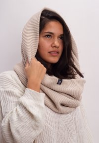 Beige knitted scarf worn as a hood, featuring a textured pattern. Accented with a small dark blue logo patch. Enveloped around the neck.