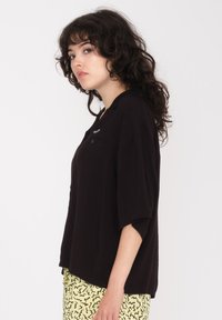 Chemise noire oversize avec une fermeture à boutons et un col. Elle possède une poche poitrine et des manches courtes, associée à une jupe jaune à motifs.