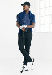 Marinblå mönstrad kortärmad piké, vit golfhandske, svarta byxor, vita sneakers med svarta ränder, håller en golfklubba, står mot en vit bakgrund.