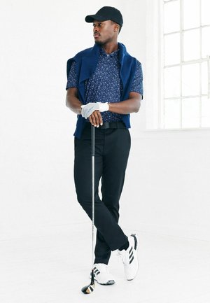 Navy īsām piedurknēm rakstaina polo krekls, balta golfa cimds, melnas bikses, baltas kedas ar melnām svītrām, turot golfa nūju, stāvot pret baltiem foniem.