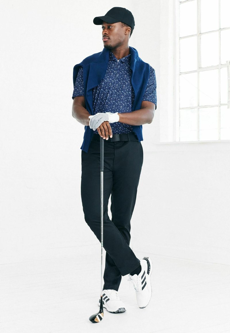Marinblå mönstrad kortärmad piké, vit golfhandske, svarta byxor, vita sneakers med svarta ränder, håller en golfklubba, står mot en vit bakgrund.