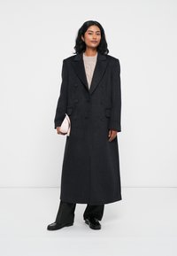Cappotto lungo doppiopetto in misto lana grigio scuro, con ampi rever e tasche laterali. Abbinato a un maglione leggero e pantaloni neri eleganti.