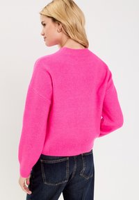 Rosa gestrickter Pullover mit rundem Ausschnitt, langen Ärmeln, geripptem Saum und weicher, flauschiger Textur. Getragen über dunklen Jeans.