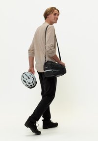 Borsa a tracolla nera con cerniere, che tiene un casco da bicicletta grigio chiaro, indossato con pantaloni neri e calzature nere. Lo sfondo è bianco uniforme.