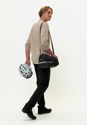 Borsa a tracolla nera con cerniere, che tiene un casco da bicicletta grigio chiaro, indossato con pantaloni neri e calzature nere. Lo sfondo è bianco uniforme.