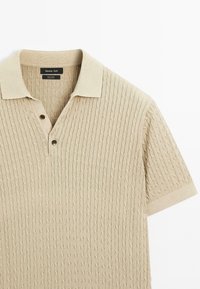 Beige strikket polo shirt lavet af 100% bomuld med et struktureret kabeldesign, korte ærmer og tre-knaps lukning.