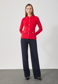 Mujer de pie con las manos detrás de la espalda, vistiendo una chaqueta roja abotonada, pantalones anchos azul marino, zapatos negros y pendientes de aro dorados.