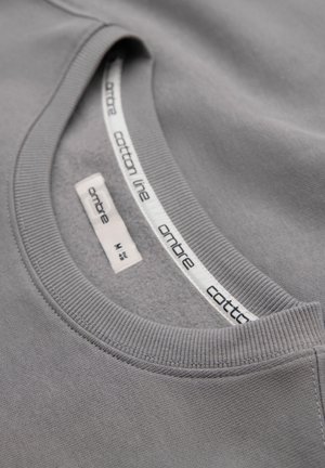Maglia grigia in cotone con scollo a girocollo che mostra le etichette interne del colletto con il nome del marchio "ambre" e la taglia "M".